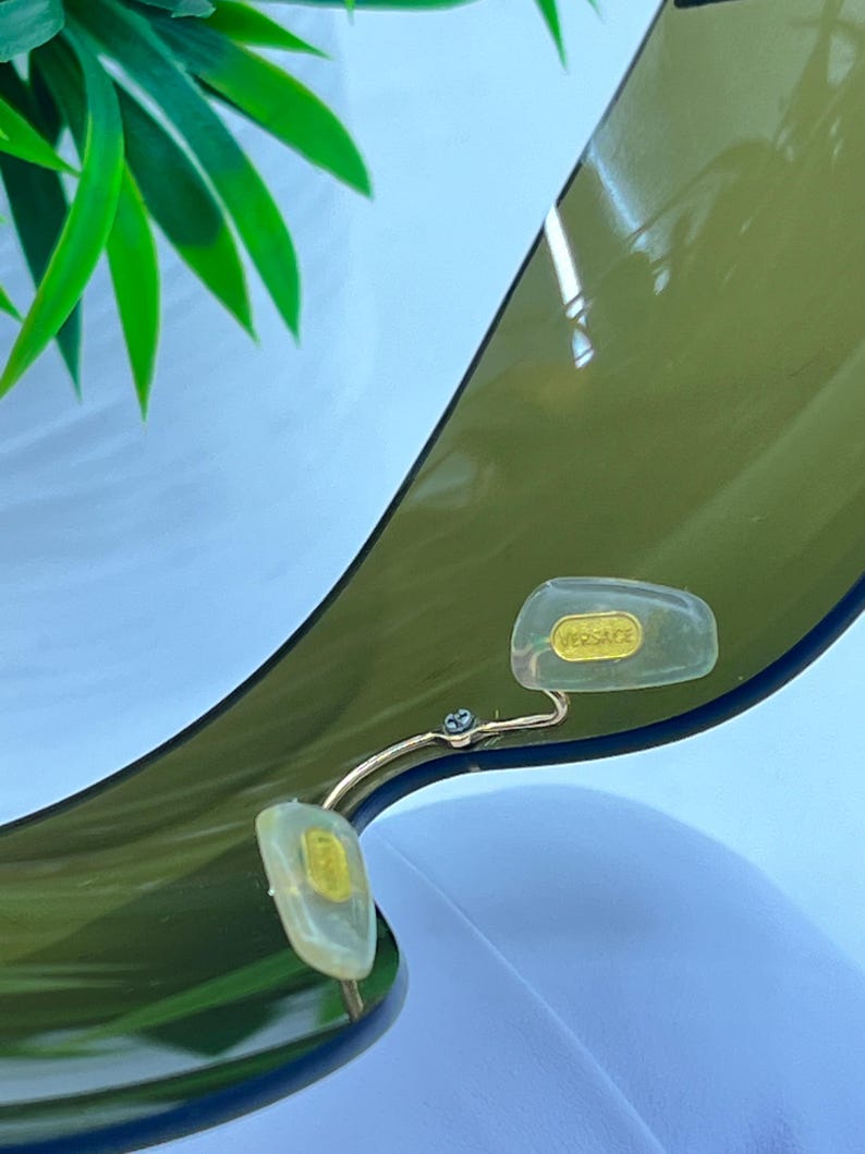 May include: A pair of olive green tinted sunglasses featuring a single continuous lens design. The nose pads are clear and translucent, with the word VERSACE embossed in gold lettering on the right nose pad, attached by a thin gold metal bridge.