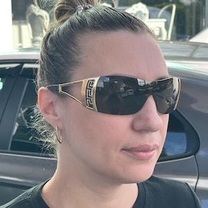 May include: Wraparound sunglasses featuring dark tinted lenses and gold-tone metal frames. The temples display a prominent gold rectangular accent with a black Greek key pattern and small embedded crystals, connecting to thin gold arms.