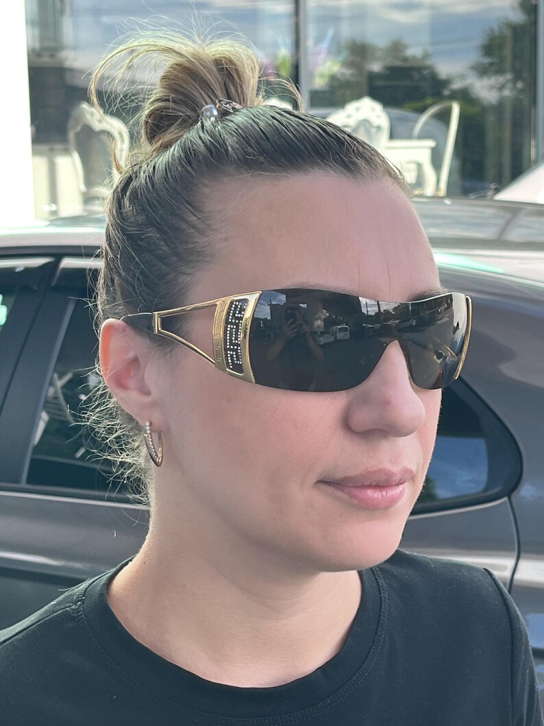 May include: Wraparound sunglasses featuring dark tinted lenses and gold-tone metal frames. The temples display a prominent gold rectangular accent with a black Greek key pattern and small embedded crystals, connecting to thin gold arms.
