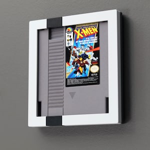 May include: Framed Nintendo Entertainment System (NES) game cartridge for "The Uncanny X-Men." The cartridge is gray, with a colorful label featuring X-Men characters. The frame is white with black accents, mounted on a gray wall.