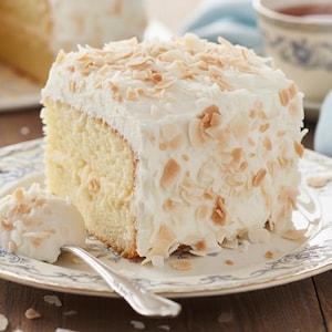 Puede incluir: Una rebanada de pastel de coco con glaseado blanco y hojuelas de coco tostadas en un plato decorativo. Una cuchara contiene una cucharada de glaseado y coco. Una taza de té y un pastel entero están en el fondo.