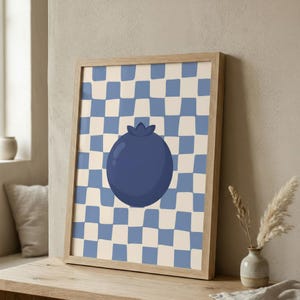 May include: A framed art print featuring a large, stylized blueberry in shades of blue. The background has a checkerboard pattern in light blue and cream. The artwork is displayed in a light wood frame, suitable for kitchen or dining room decor.