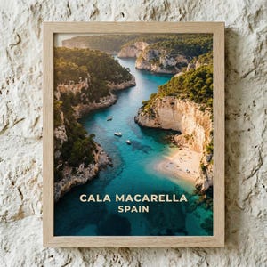 May include: Framed print of Cala Macarella beach in Spain. The image shows a turquoise bay surrounded by cliffs and lush greenery. Several boats are in the water, and the text "CALA MACARELLA SPAIN" is at the bottom.