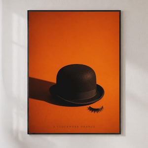 May include: A framed poster featuring a black bowler hat and a single set of false eyelashes on a solid orange background. The text A CLOCKWORK ORANGE is printed in small black capital letters at the bottom center of the composition.