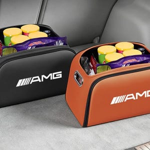 Könnte beinhalten: Zwei rechteckige Aufbewahrungsboxen, eine in Schwarz und eine in Orange, mit dem weißen AMG-Logo auf der Vorderseite. Jeder Behälter verfügt über eine Reißverschlussöffnung an der Oberseite, seitliche Griffe und ist mit Lebensmitteln gefüllt.