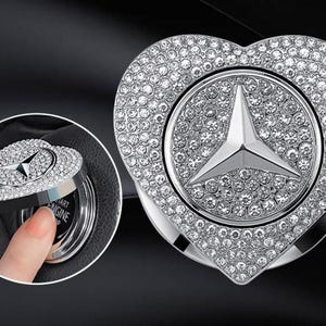 Op de afbeelding: Een hartvormige startknopafdekking voor een auto met een zilverkleurige afwerking en ingebedde heldere kristallen. Het midden toont een opvallend driepuntig sterembleem. Het accessoire is ontworpen om over een ronde ontstekingsknop te passen.