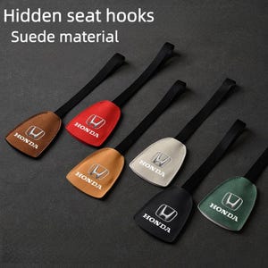 Puede incluir: Seis ganchos de asiento ocultos de material de ante con correas negras, cada uno con un logotipo de Honda plateado y el texto Honda. Los ganchos vienen en colores marrón, rojo, tostado, beige, negro y verde, dispuestos en diagonal sobre una superficie oscura.