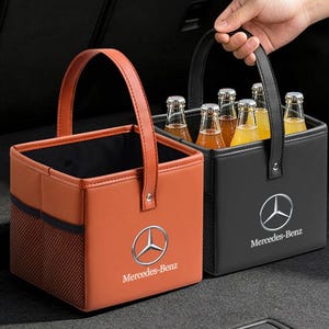 Könnte beinhalten: Zwei quadratische Aufbewahrungskörbe mit Griffen, einer in Orange und einer in Schwarz, beide mit dem Mercedes-Benz Logo und Schriftzug. Der schwarze Korb enthält sechs Glasflaschen mit bernsteinfarbener Flüssigkeit. Jeder Korb hat seitliche Netztaschen.