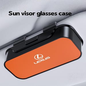 May include: A black and orange car sun visor glasses case featuring the Lexus logo in white. The rectangular accessory is attached to a light gray vehicle sun visor and includes the text Sun visor glasses case printed above the item.