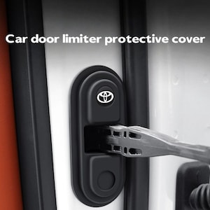 May include: A black, oval-shaped car door limiter protective cover installed on a white vehicle door frame. The cover features a white Toyota logo at the top and includes the text Car door limiter protective cover printed above the component.