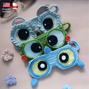 May include: Three crocheted eye masks featuring large, round, dark eyes and small ears. The top mask is light blue, the middle is green with a small red accent, and the bottom is light blue with yellow-green eye circles. Each has a metal ring attached.