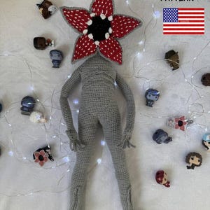 May include: A crocheted Demogorgon doll with a red and white flower-like head, gray body, and long limbs. The doll is surrounded by small character figurines and string lights. The text "CROCHET PATTERN" and a US flag are visible.