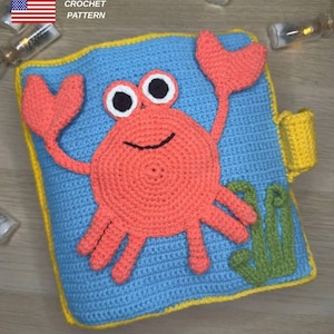 Op de afbeelding: Gehaakt babyboekje met een krabthema, met een blauwe achtergrond en gele rand. De krab is oranje met zwart-witte ogen. Het boek heeft een klein geel zakje en groene zeewieraccenten. De tekst "CROCHET PATTERN" is zichtbaar.