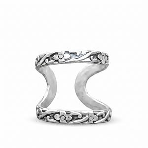 May include: A silver-colored double-band ring with a floral design. The ring features two bands connected by curved supports. The bands are adorned with detailed floral patterns, creating an elegant and nature-inspired aesthetic. The ring is set against a white background.