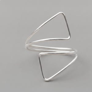 May include: A silver-toned metal ring featuring a continuous, open-ended design that forms two distinct, stacked triangular shapes. The smooth, polished wire construction wraps around to create an adjustable, geometric silhouette with clean lines.