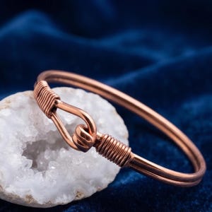 Puede incluir: Una pulsera de alambre de cobre con un mecanismo de cierre de gancho y ojo. La pulsera está construida con dos hilos paralelos de alambre de cobre, con cada extremo envuelto en una bobina apretada para formar el cierre. El metal tiene un acabado.