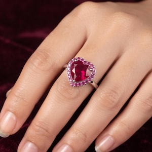 Pode incluir: Um anel de tom prateado com uma grande pedra preciosa vermelho-escura em forma de coração no centro. A pedra central é rodeada por um halo de pedras mais pequenas, redondas e cor-de-rosa brilhante, montadas numa banda prateada polida e simples.
