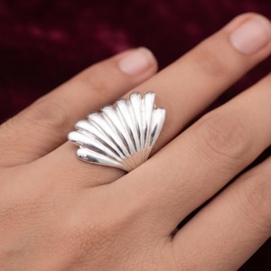 May include: A polished silver ring featuring a fan-shaped design with multiple raised, vertical ridges that mimic the appearance of a seashell. The metallic surface reflects light, highlighting the curved, radiating segments of the ring.