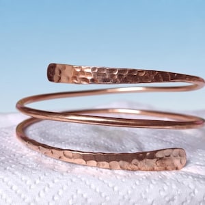 May include: A copper spiral bracelet featuring a continuous coil design. The metal surface displays a distinct hammered texture on the tapered ends, while the central portion of the wire remains smooth and rounded, creating a metallic finish.