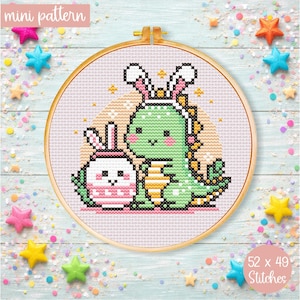 May include: A cross-stitch pattern in a wooden hoop features a green dinosaur and a pink bunny, both wearing bunny ears. The text "mini pattern" is at the top, and "52 x 49 Stitches" is at the bottom. The background is light pink.