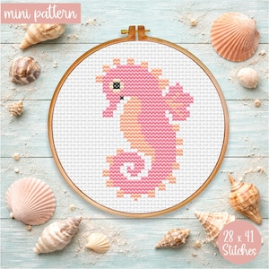 May include: A cross-stitch pattern of a pink seahorse inside a wooden embroidery hoop. The design is surrounded by seashells on a light blue wooden surface. The text "mini pattern" and "28 x 41 Stitches" are visible.