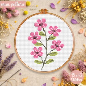 May include: A cross-stitch floral pattern in a wooden embroidery hoop. The design features pink flowers with dark centers and green leaves on a black stem. The text "mini pattern" is at the top, and "45 x 63 Stitches" is at the bottom.