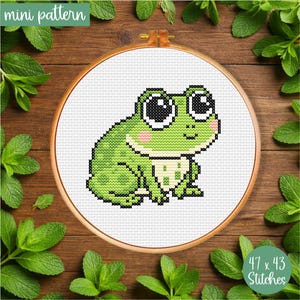 May include: A cross-stitch pattern of a green frog with large black and white eyes, pink cheeks, and a white belly, set in a wooden embroidery hoop. The text "mini pattern" and "47 x 43 Stitches" are visible.