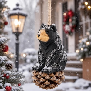 May include: A hand-carved wooden bear ornament with a black body and a light tan face, perched atop a natural brown pinecone. The bear features small black eyes and is suspended by a rustic jute twine string, lightly dusted with white snow.