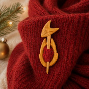 May include: A light brown wooden shawl pin featuring a stylized fox head design with a pointed top piece and a rounded base piece, securing a section of deep red knitted fabric. The wooden accessory has a smooth finish and visible wood grain.