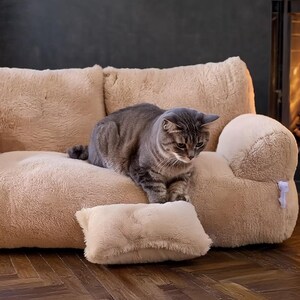 May include: A plush, beige cat sofa with a matching pillow. A gray tabby cat sits on the sofa, looking down. A fireplace with a burning fire is in the background, creating a cozy atmosphere.