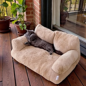 May include: A gray cat lounges on a beige pet sofa on a wooden deck. The sofa has a textured, plush appearance with rounded armrests. Potted plants are visible in the background, creating a relaxed outdoor setting. The cat is the focal point, enjoying the comfort of the sofa.