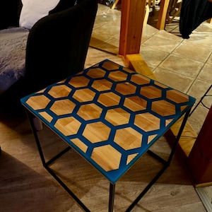 Könnte beinhalten: Quadratischer Beistelltisch mit geometrischem Wabenmuster. Die Tischplatte zeigt sechseckige Holzformen, die in blauem Harz eingelassen sind, getragen von einem schwarzen Metallrahmen. Der Tisch ist ca. 46 cm breit und 46 cm lang.