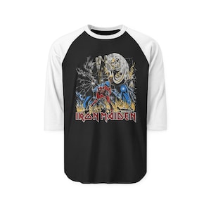 Puede incluir: Camiseta de béisbol raglán en blanco y negro con un gráfico de un monstruo y una figura en rojo y azul, con el texto "IRON MAIDEN" en rojo y blanco. El diseño está sobre un fondo de llamas y relámpagos.
