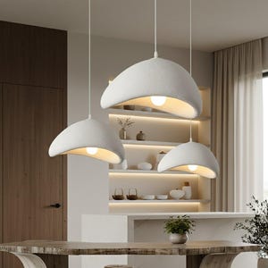 Może przedstawiać: Trzy białe, teksturowane lampy wiszące o zakrzywionym, organicznym kształcie zwisają z sufitu. Każda lampa ma ciepłą, świecącą żarówkę. Lampy zawieszone są nad drewnianym stołem w nowoczesnej kuchni.