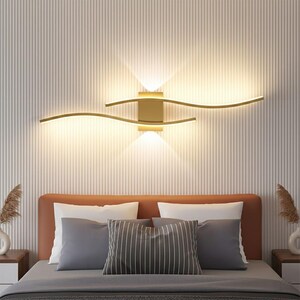 Può includere: Un'applique da parete moderna color oro con un design unico e fluente. L'apparecchio ha due bracci curvi che si estendono da una base quadrata centrale, emettendo un bagliore caldo. L'applique è montata su una parete a strisce verticali sopra un letto.