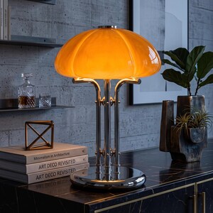 Peut inclure: Une lampe de table avec un abat-jour en verre orange arrondi et lumineux, reposant sur une base en métal chromé poli dotée de trois tiges de support verticales. La lampe est fixée sur un socle circulaire chromé et diffuse une lumière chaude.