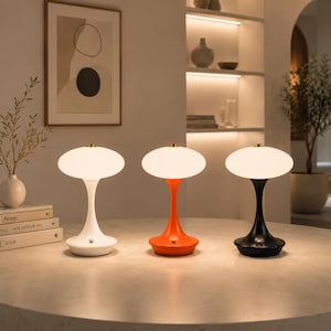 May include: Three table lamps with mushroom-shaped white glass shades and smooth, curved bases in white, orange, and black. Each lamp features a small metallic button on the base and emits a soft, warm glow from the rounded, translucent glass top.