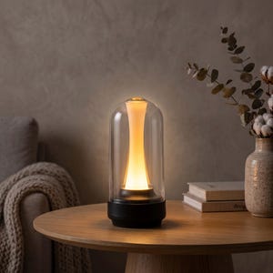 Könnte beinhalten: Eine zylindrische Tischleuchte mit einer klaren Glaskuppel über einer zentralen, sanduhrförmigen Lichtquelle, die ein warmes, sanftes Licht ausstrahlt. Die Leuchte steht auf einem soliden, schwarzen runden Sockel für einen stabilen Halt.