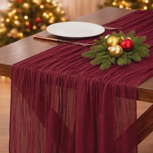 May include: A burgundy table runner draped over a wooden table, with a white plate and a knife. A small Christmas decoration with gold and red ornaments and green pine branches sits on the table. A Christmas tree is in the background.