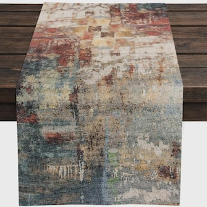 May include: Abstract patterned table runner in shades of red, blue, beige, and brown. The rectangular runner features a textured design with a blend of colors, creating a visual effect. The runner is placed on a dark wooden surface.
