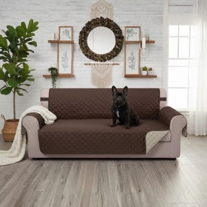May include: A brown quilted sofa cover with a black dog sitting on it. The sofa has light beige arms and a white blanket draped over one arm. The background includes a white brick wall, a mirror, and framed artwork.