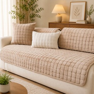 May include: A beige sofa cover with a raised square grid texture, featuring a matching square throw pillow and a rectangular white throw pillow. The cover drapes over the seat and backrest of the sofa, providing a uniform, quilted appearance.