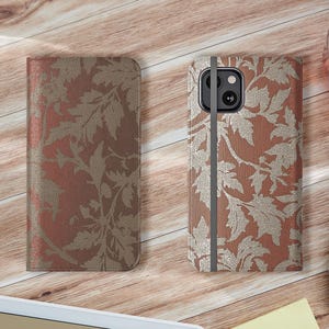 May include: Two phone cases with a floral design. One is closed, the other open, showing a phone. The cases are a reddish-brown color with a gold leaf pattern. The cases are on a wooden surface.
