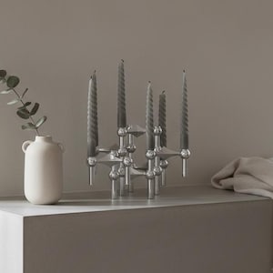 May include: A modern interior scene featuring a white vase with a sprig of greenery, a silver candelabra with gray candles, and a folded beige blanket. The candelabra holds five gray spiral candles. The objects are on a white surface.