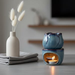 May include: A blue ceramic essential oil diffuser in the shape of a tulip, with a lit tea light inside. A small white vase with dried flowers and a folded gray cloth are also in the image.