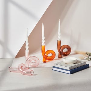 May include: A selection of decorative glass candle holders in pink, orange, and brown, with white spiral candles. A small white bowl sits on a stack of books. The candle holders have a unique, curved design.