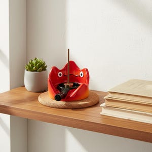 Op de afbeelding: Een rood en oranje keramische wierookhouder met een cartoonachtig gezichtsontwerp. De houder heeft een zwarte opening voor wierook en een goudkleurige metalen wierookstokhouder. Hij staat op een kurken onderzetter op een houten plank, met een kleine vetplant en boeken in de buurt.