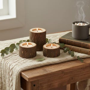 Op de afbeelding: Drie houten kandelaars met brandende kaarsen van verschillende groottes staan op een houten tafel. Een grijze mok met marshmallows en stoom staat op boeken. Eucalyptustakken en een crèmekleurige gebreide loper maken de scène compleet.