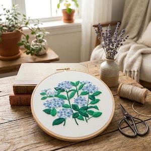 Puede incluir: Un bastidor de bordado redondo con un diseño floral detallado de flores de hortensia azules y hojas verdes. El bordado está sobre una tela blanca, enmarcado por un aro de madera natural. La escena incluye un jarrón con lavanda y decoración vintage.