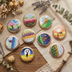 May include: A selection of round compact mirrors, each with an embroidered landscape design. The mirrors depict various scenes, such as a road with trees, a field of flowers, mountains, a desert with a camel, and a deer under a full moon. They have gold or copper-coloured frames.